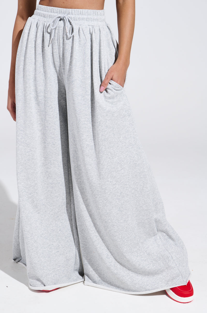Styled in COZY AND COMFY SWEATPANT with a wide-leg silhouette and adjustable drawstring waist, a person stands with one hand in their pocket. Red shoes peek out from beneath the sweeping pants' legs against a plain white background.