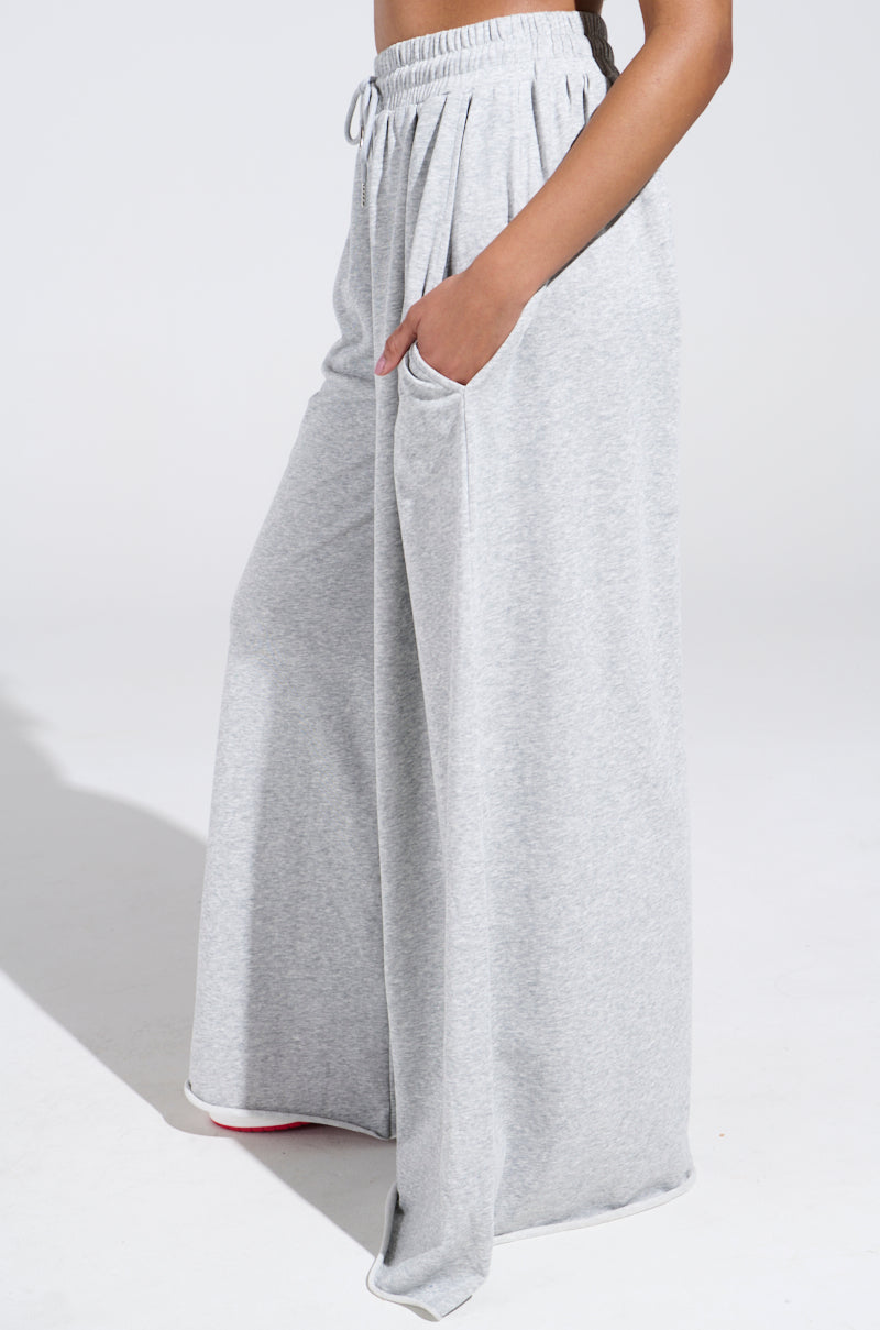 A person models the COZY AND COMFY SWEATPANT, featuring a high-rise, wide-leg silhouette in light gray. Their hands are tucked in pockets as the plain white backdrop accentuates the casual elegance of their attire.