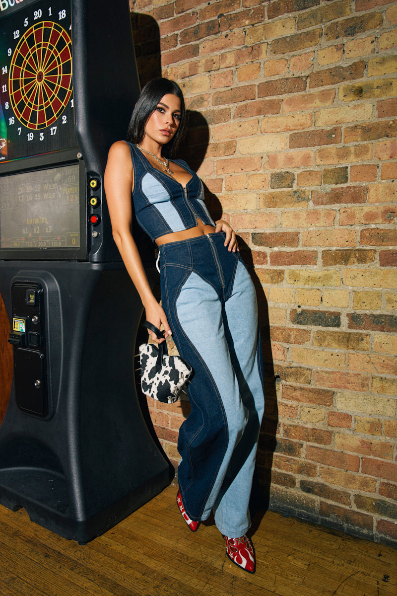 A woman poses indoors near a dartboard, wearing FIRST CLASS RELAXED DENIM PANT, a two-tone denim patchwork crop top, red shoes, and holding a black-and-white patterned handbag.