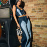 A woman poses indoors near a dartboard, wearing FIRST CLASS RELAXED DENIM PANT, a two-tone denim patchwork crop top, red shoes, and holding a black-and-white patterned handbag.