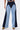A person wears the FIRST CLASS RELAXED DENIM PANT—high-waisted, two-tone wide-leg jeans with a front zipper—and yellow patterned shoes. Their upper body is partially visible.