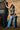 A woman in a two-tone denim patchwork top and FIRST CLASS RELAXED DENIM PANT poses by a dartboard machine against a brick wall, holding a black-and-white cow print bag and wearing red patterned shoes for a statement look.