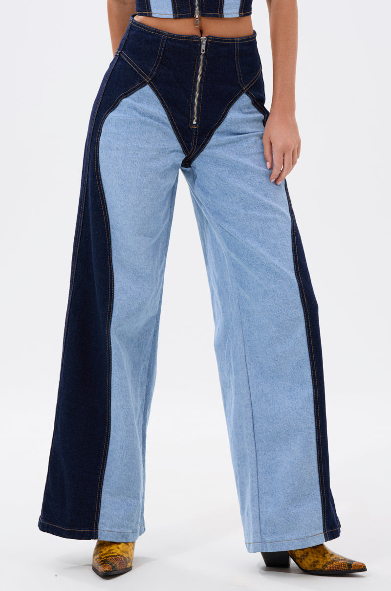 A person wears the FIRST CLASS RELAXED DENIM PANT featuring a wide-leg silhouette and bold two-tone panels, paired with yellow snake-patterned shoes; torso partially seen against a white background.