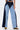 A person wears the FIRST CLASS RELAXED DENIM PANT featuring a wide-leg silhouette and bold two-tone panels, paired with yellow snake-patterned shoes; torso partially seen against a white background.
