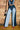 A person stands against a brick wall, wearing a blue denim corset top and the FIRST CLASS RELAXED DENIM PANT, paired with red flame-patterned shoes and holding a small black and white bag.
