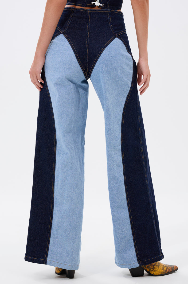 A person seen from the back wears FIRST CLASS RELAXED DENIM PANT, a high-waisted, two-tone wide-leg jean with dark and light blue panels, bold angular contrast stitching, paired with brown boots.
