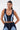 A woman with straight, dark hair and large hoop earrings wears the FIRST CLASS DENIM CORSET, a zip-front contrast-paneled crop top, with matching high-waisted denim pants. She stands with her hands behind her back.