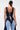 A woman with long dark hair in a ponytail models the FIRST CLASS DENIM CORSET, a lace-up back denim crop top with matching contrast-paneled cropped pants, shown from behind against a plain background.