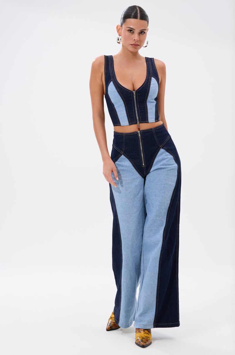 A woman models the FIRST CLASS DENIM CORSET, a sleeveless zip-up crop top featuring contrasting dark and light blue denim panels, paired with matching high-waisted wide-leg jeans. She accessorizes with hoop earrings and yellow patterned shoes.