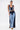 A woman models the FIRST CLASS DENIM CORSET, a sleeveless zip-up crop top featuring contrasting dark and light blue denim panels, paired with matching high-waisted wide-leg jeans. She accessorizes with hoop earrings and yellow patterned shoes.