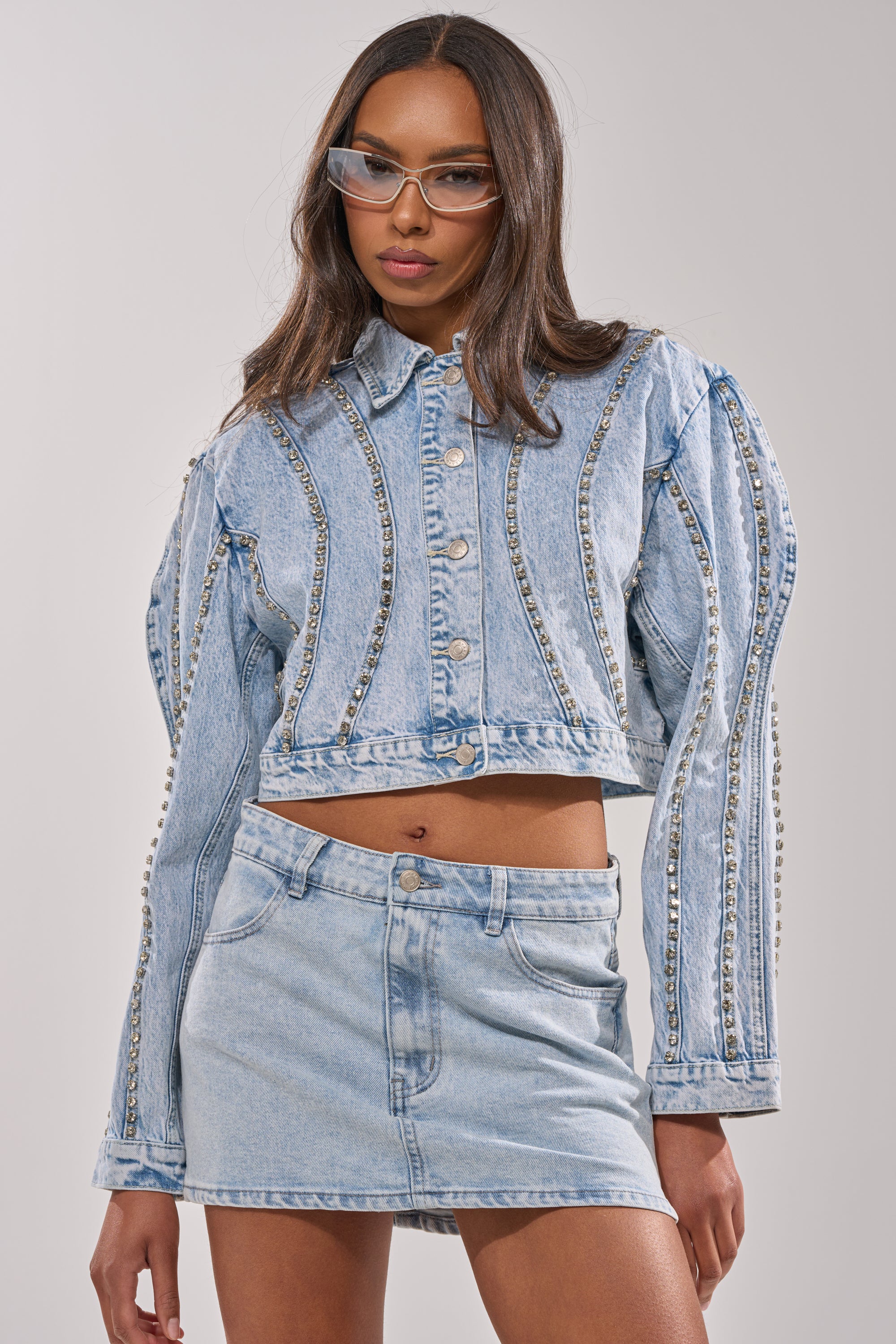 A woman wears the EXTRA EXTRA EMBELLISHED DENIM JACKET IN LIGHT BLUE, paired with a matching denim skirt and clear-frame glasses, posing confidently against a simple background. 0