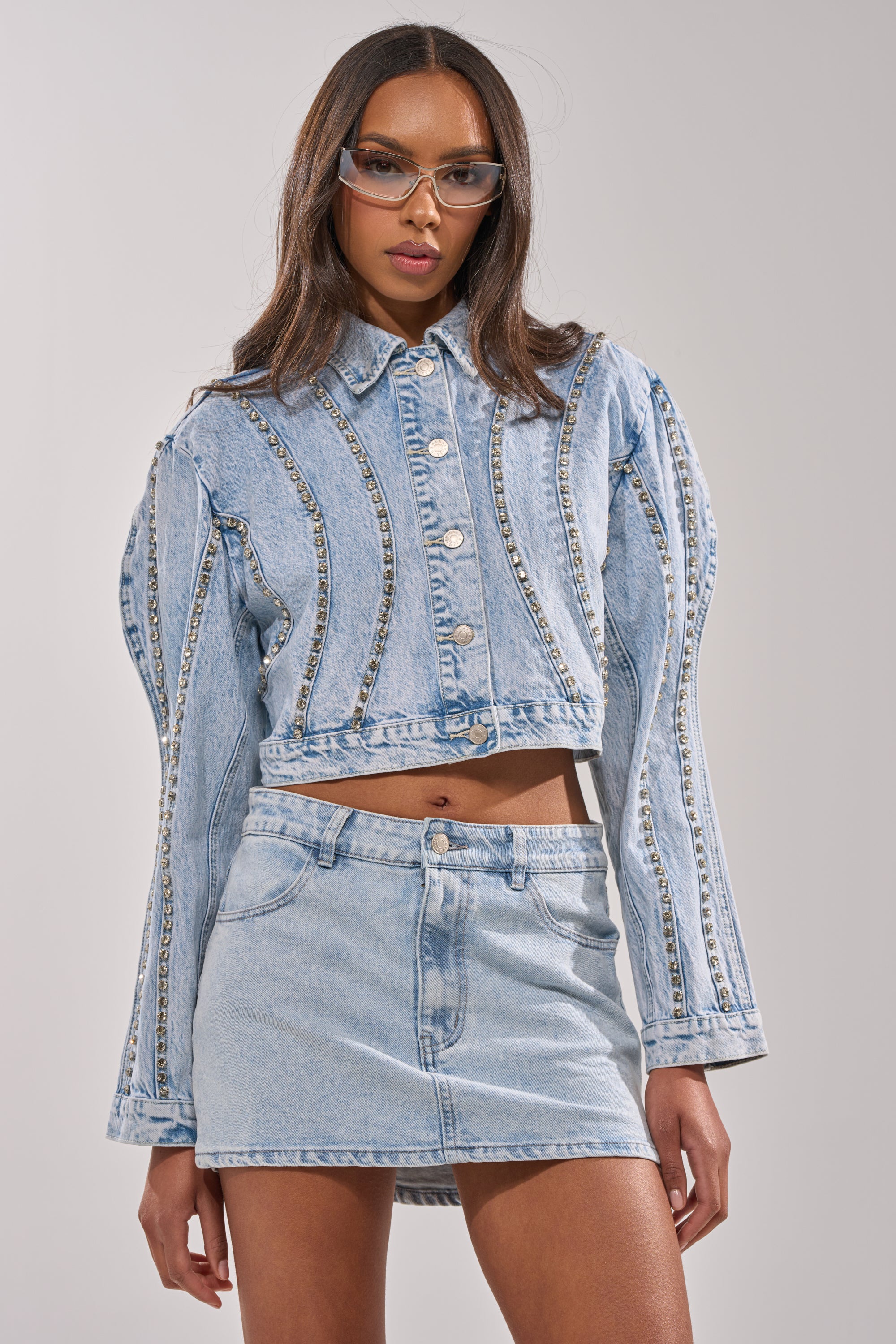 A woman wearing clear glasses and the EXTRA EXTRA EMBELLISHED DENIM JACKET IN LIGHT BLUE styled with a matching light blue denim mini skirt stands against a plain background. 1