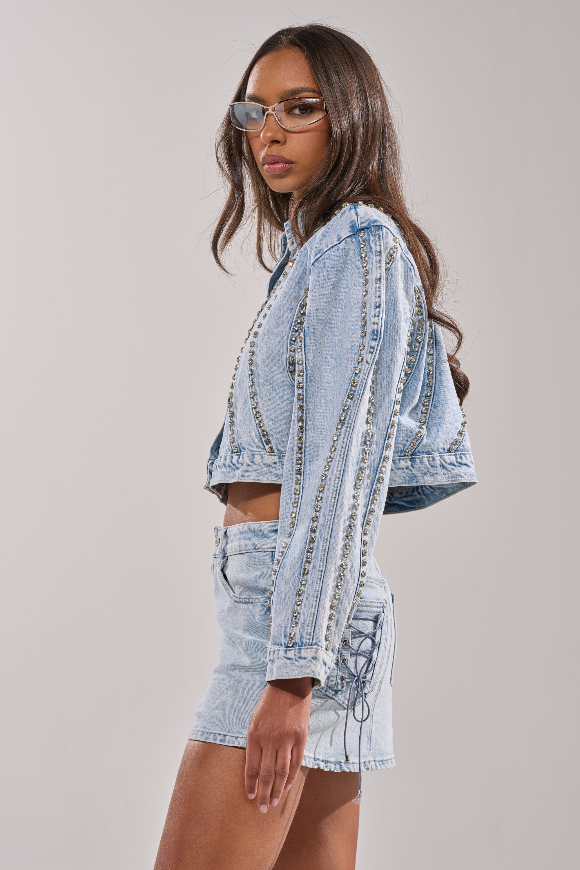 A woman with long brown hair and clear glasses wears the EXTRA EXTRA EMBELLISHED DENIM JACKET IN LIGHT BLUE, styled with matching high-waisted shorts, standing against a plain light background. 2