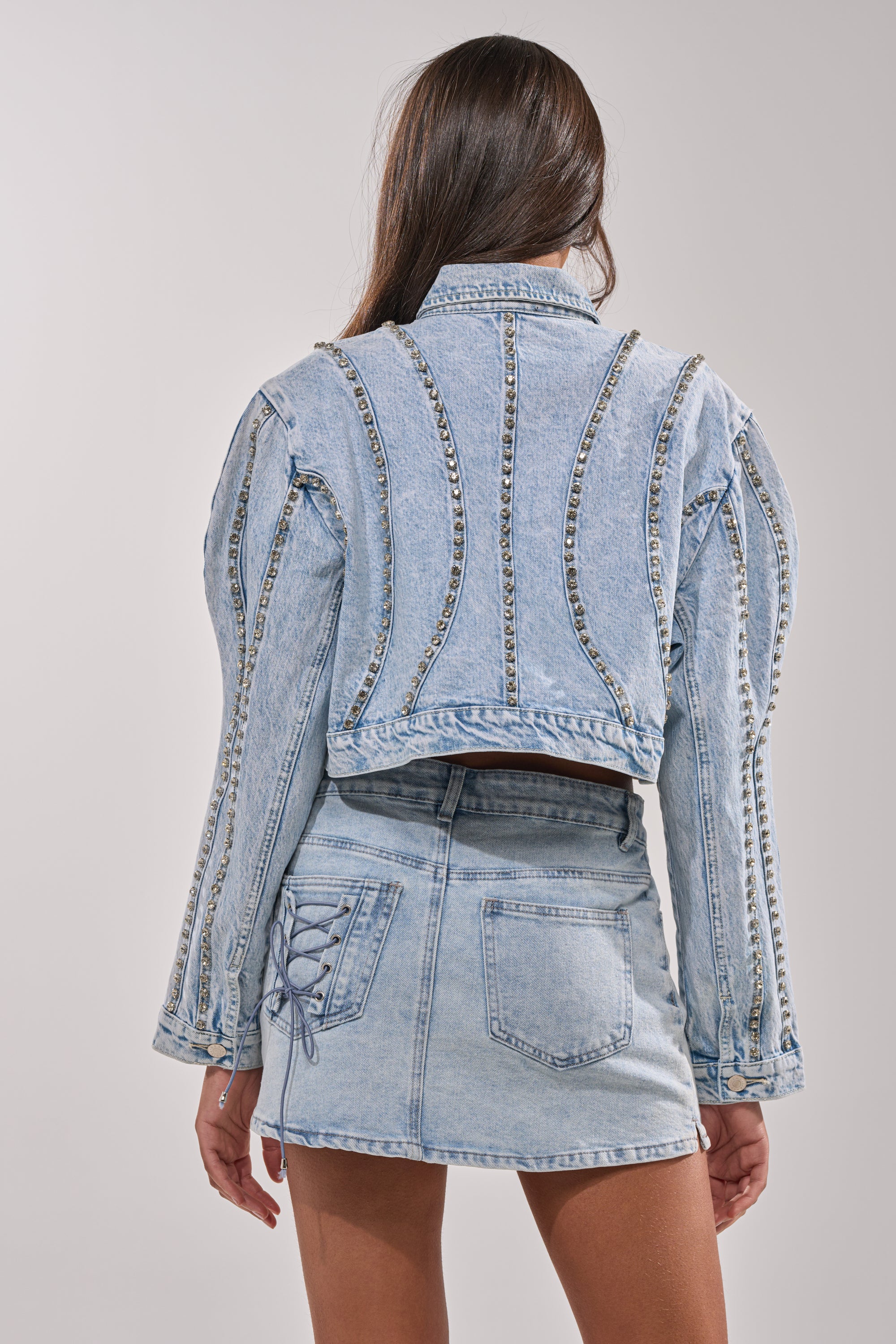 A woman faces away from the camera in the EXTRA EXTRA EMBELLISHED DENIM JACKET IN LIGHT BLUE, featuring sparkling silver details, paired with a matching lace-up side denim mini skirt. 3