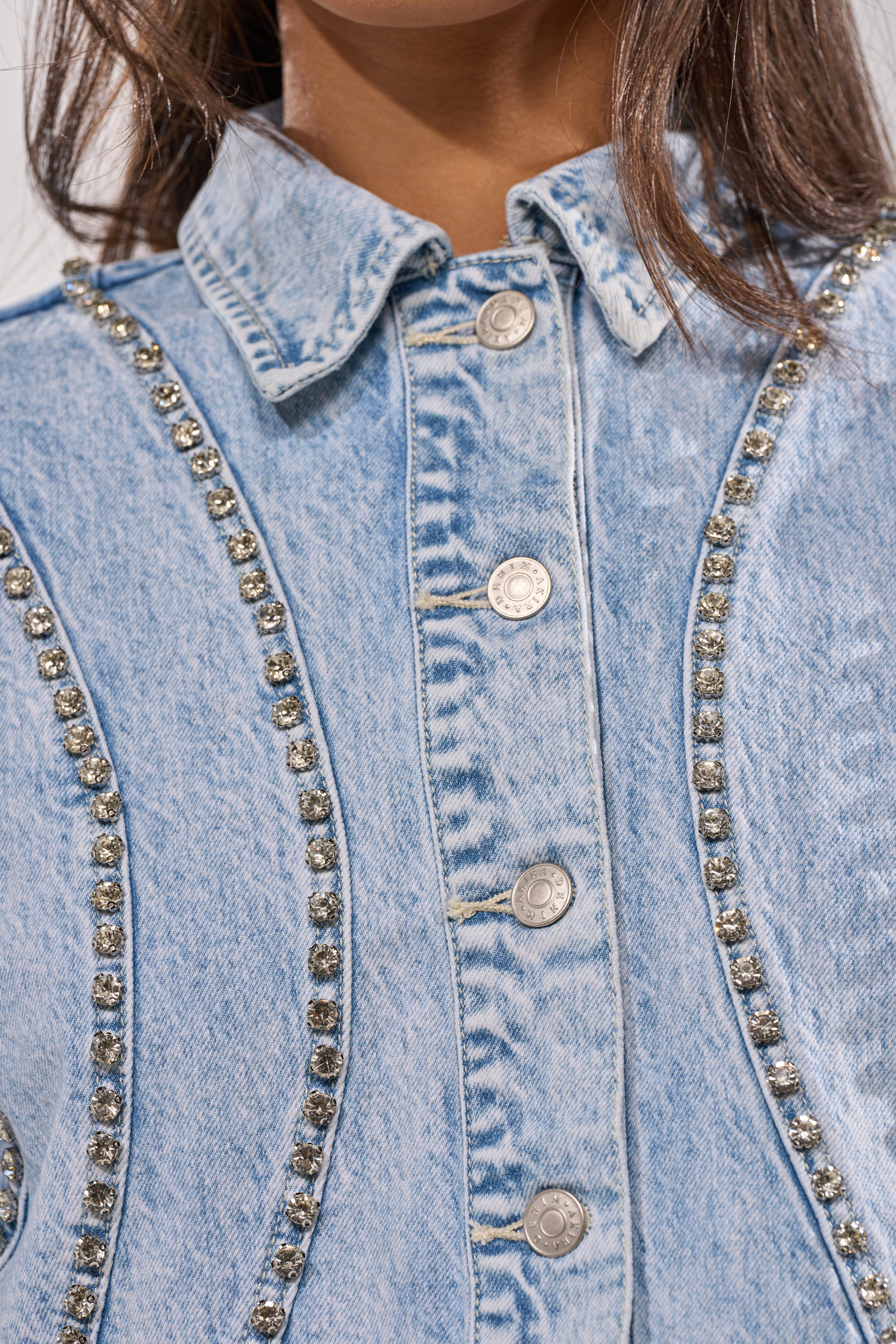 Close-up of a person wearing the EXTRA EXTRA EMBELLISHED DENIM JACKET IN LIGHT BLUE, featuring silver buttons and rhinestone trim on the seams. The collar is partially covered by the person's hair. 5