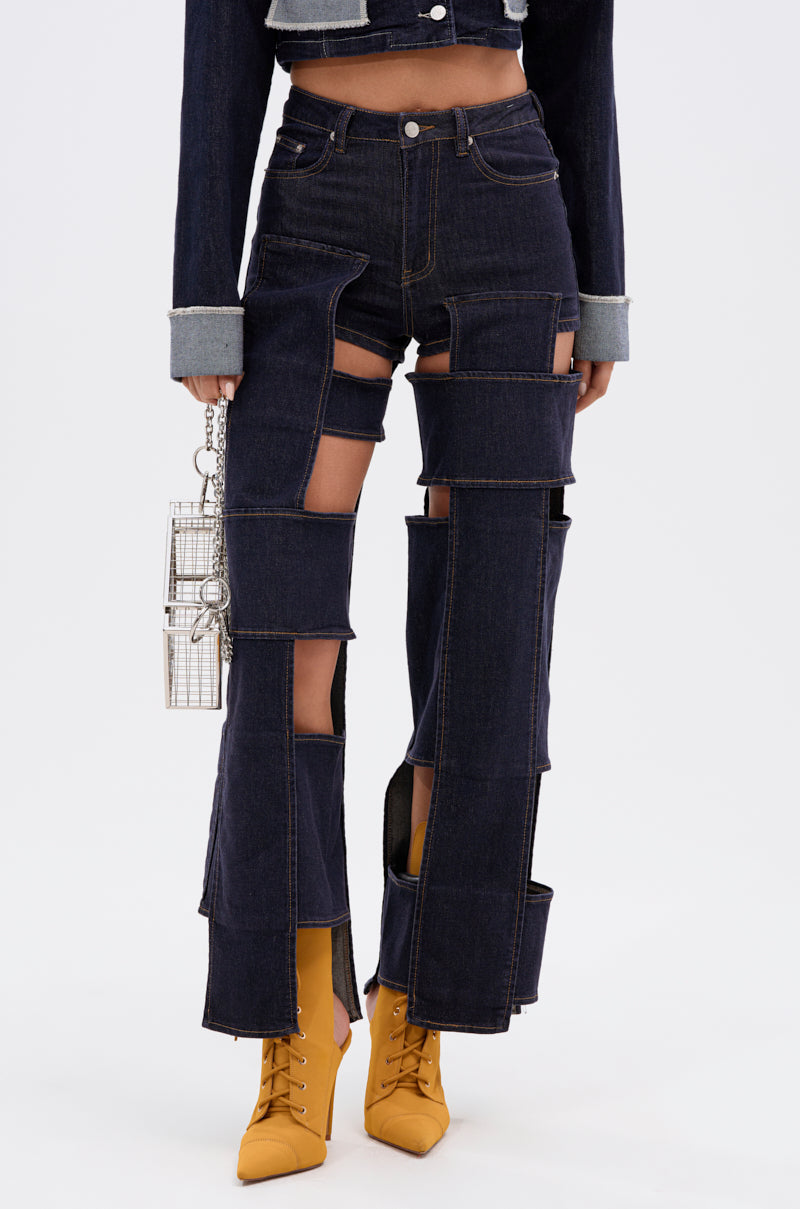 A person wears the LOCKED UP FOR GOOD DENIM PANT, featuring dark blue high-rise and cut-out designs, with a matching denim jacket, a silver purse, and yellow lace-up boots against a plain white background.