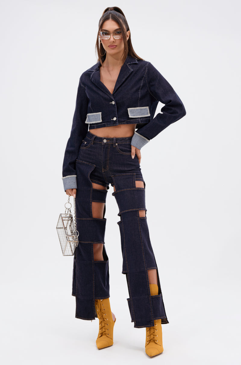 A woman showcases the LOCKED UP FOR GOOD DENIM PANT, a unique wide-leg cut-out jean, paired with a cropped denim jacket. She completes the look with yellow boots, oversized glasses, and a geometric clear handbag.