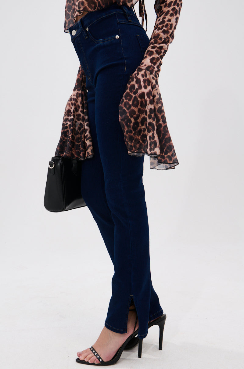 Wearing THE BEST CORSET WAISTED FLARE DENIM with small ankle slits, a sheer leopard print top with flared sleeves, black high-heeled sandals, and a black handbag, the person stands against a plain white background.