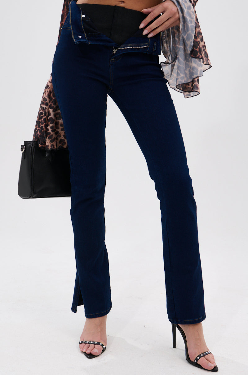 Wearing THE BEST CORSET WAISTED FLARE DENIM, unzipped, with black sandals, a sheer animal print top, and holding a black handbag, a person stands against a plain white background.