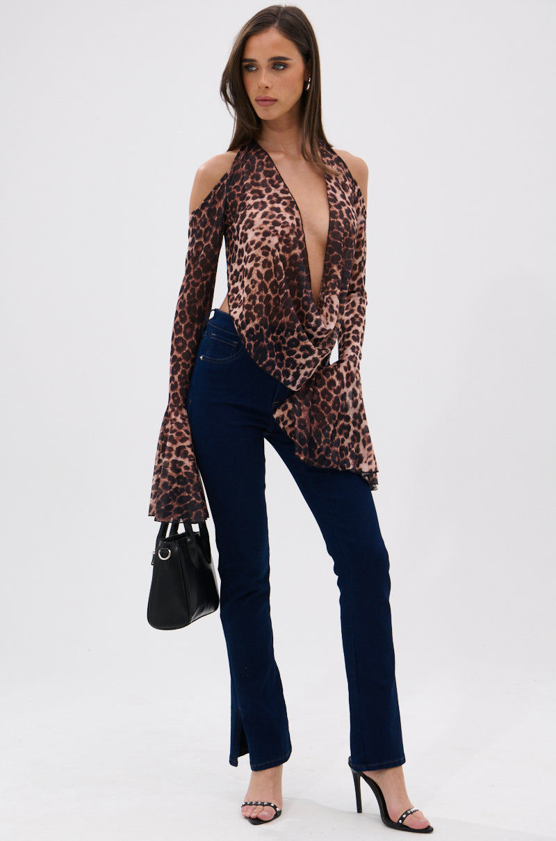 A woman in a deep V-neck, long-sleeve leopard print blouse with cold shoulders, THE BEST CORSET WAISTED FLARE DENIM, black heels, and a black handbag stands against a white background.