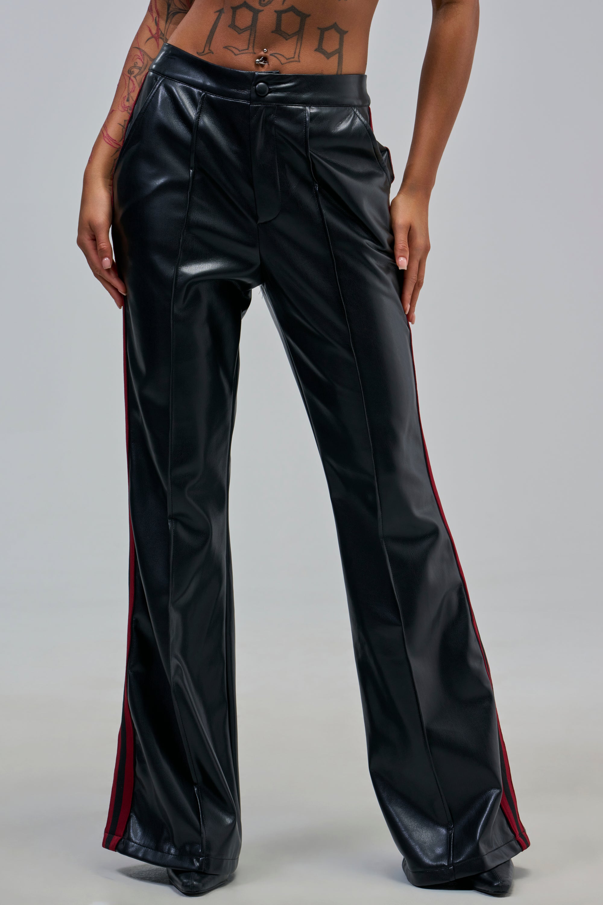 A person wearing COME AND GET IT TROUSER with black faux leather, red side stripes, and flared legs stands hands on hips, displaying tattoos on their torso and arm.