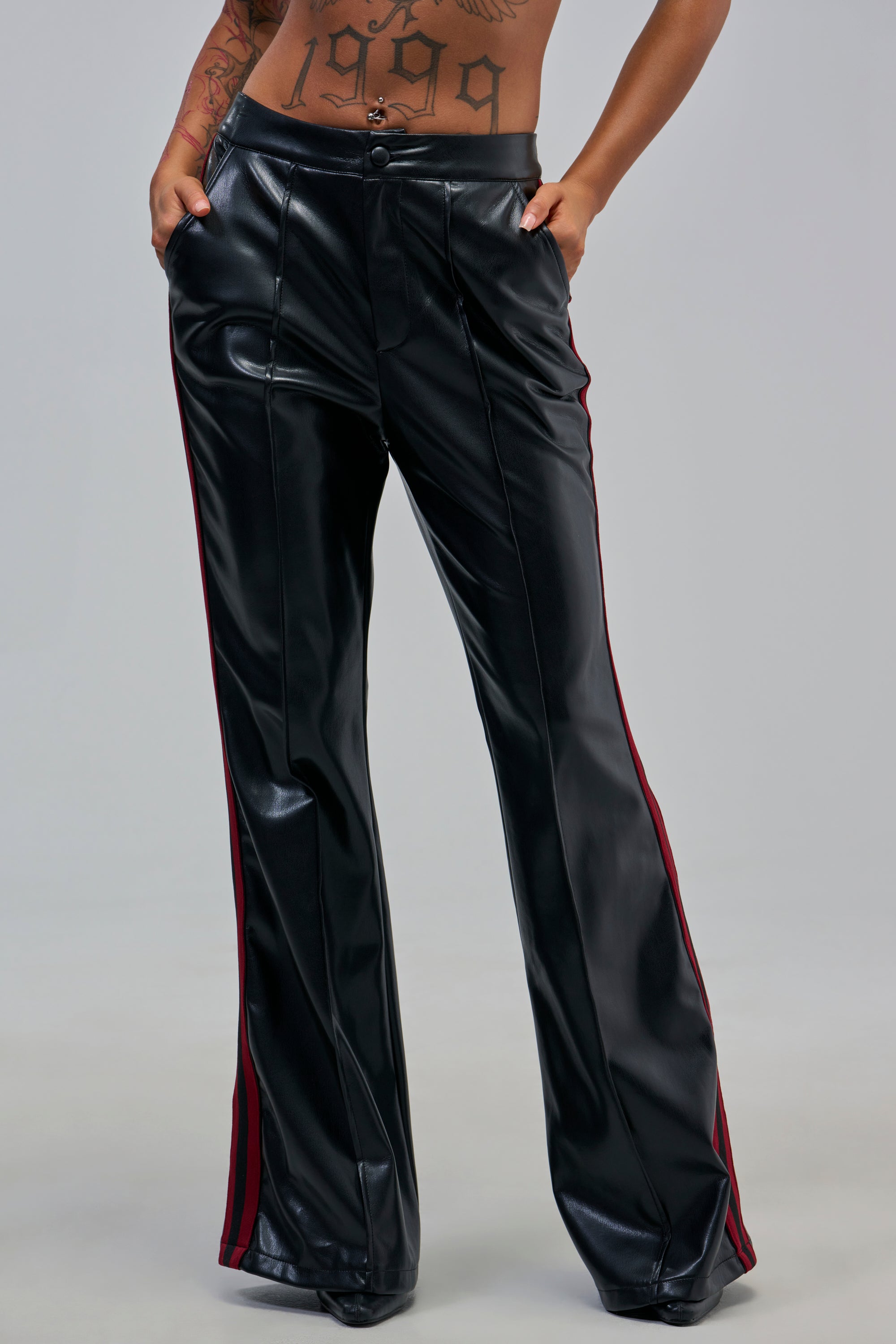 Model in COME AND GET IT TROUSER—black faux leather with red side stripes, high-rise waist, flared legs. Hands in pockets show midsection tattoos. Plain gray background.