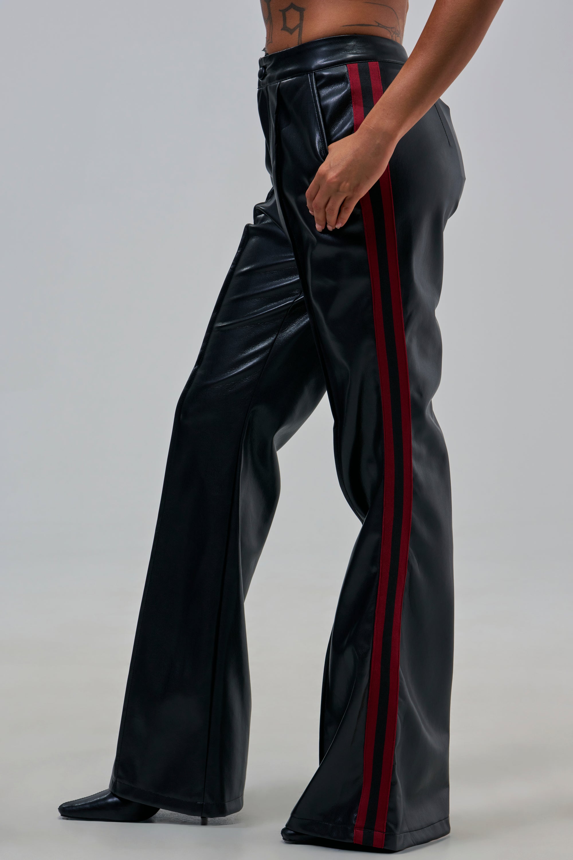 Model wears the COME AND GET IT TROUSER—black faux leather flares with red side stripes and a high-rise fit—against a light gray background. Only the lower body and a torso star tattoo are visible.