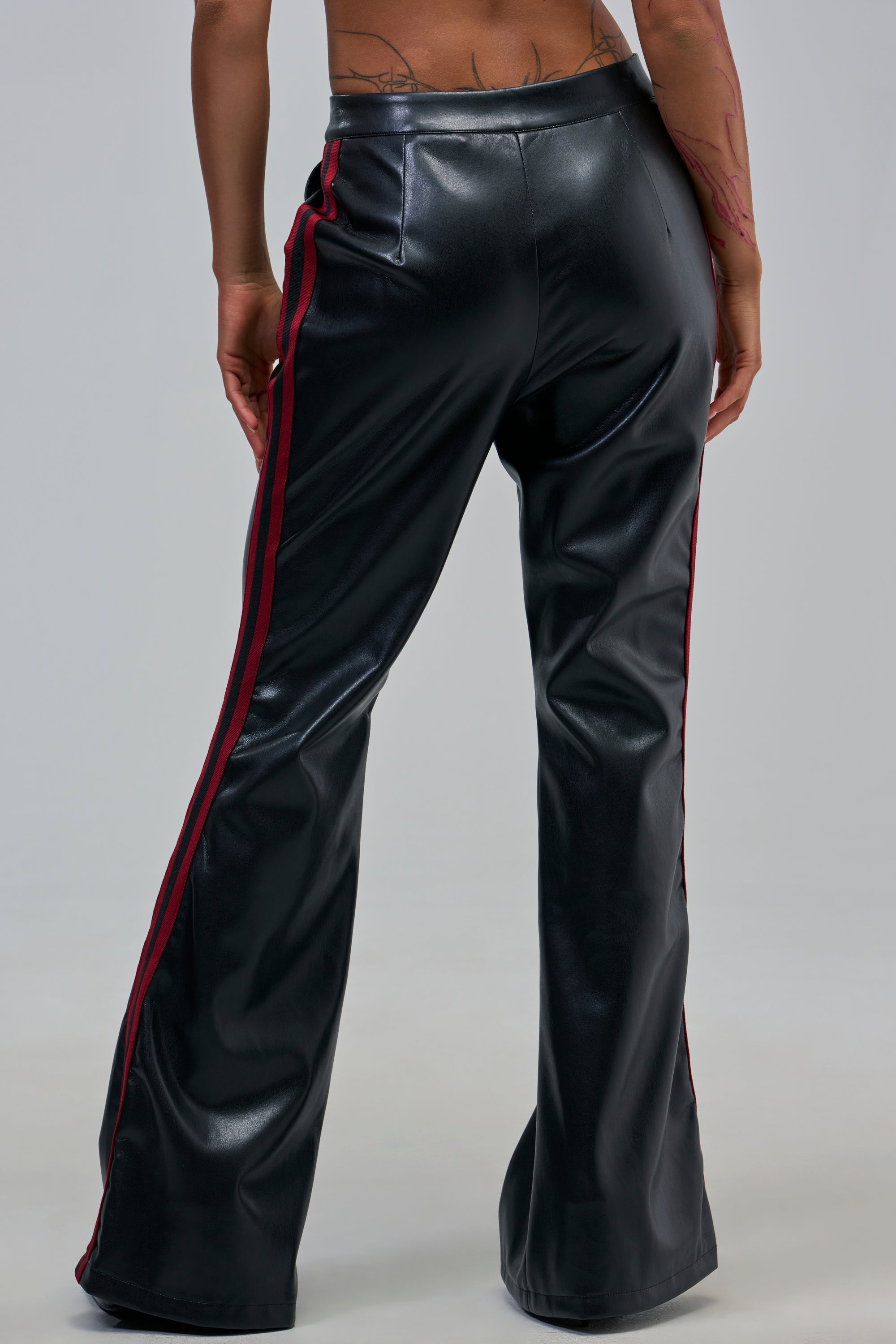 Person shown from the back wearing COME AND GET IT TROUSER—black faux leather, flared-leg, high-rise fit with red side stripes and a shiny finish, set against a plain gray background.