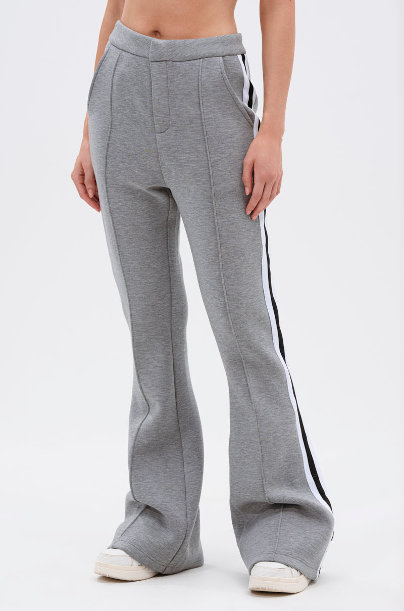 Wearing the YOU KNOW THE DRILL TROUSER, a high-rise gray flared pant with vertical seams and black-and-white side stripes, paired with white sneakers. The photo is cropped at the midriff and ankles for a retro vibe.
