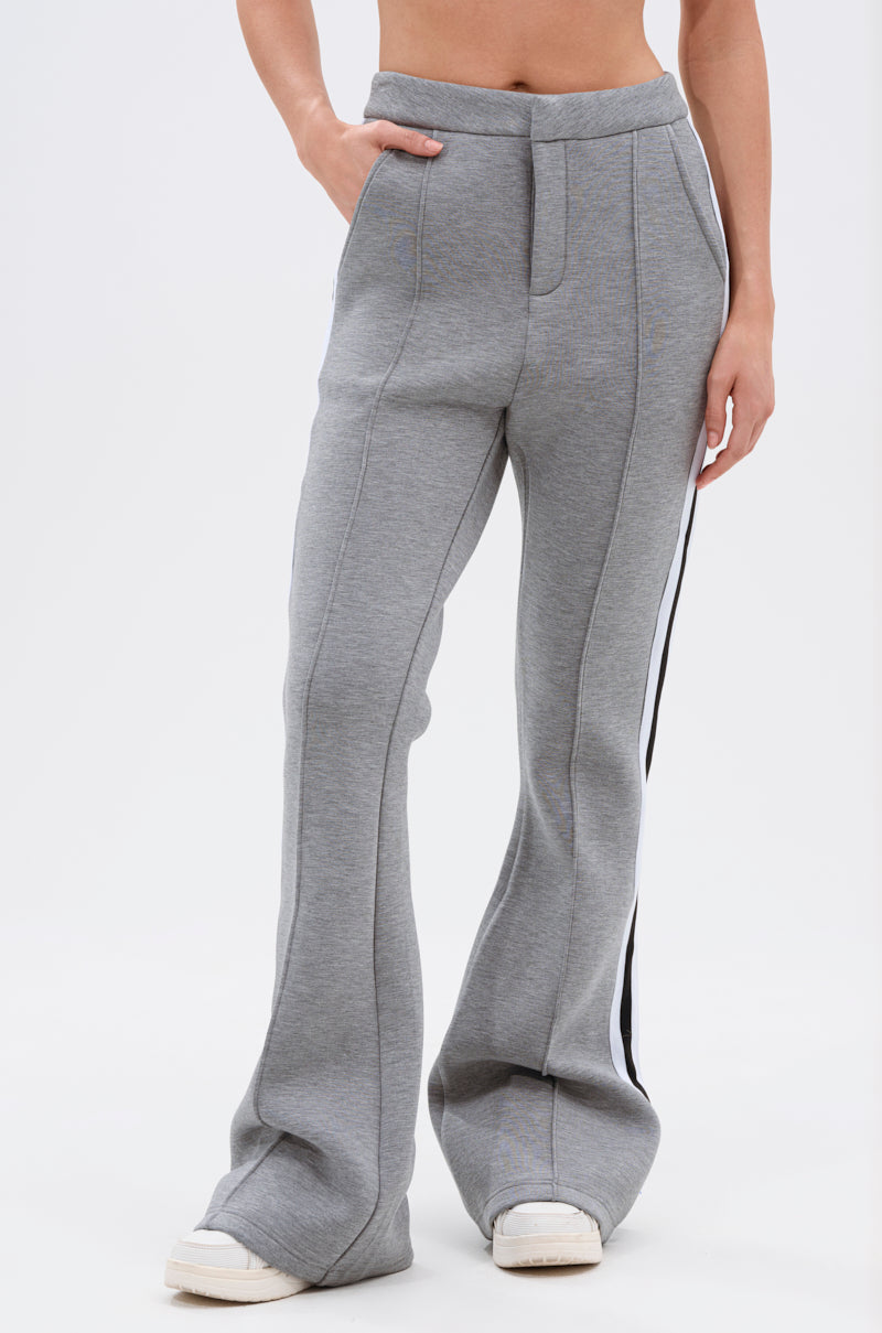 Someone models the YOU KNOW THE DRILL TROUSER—high-waisted, gray flared athleisure pants featuring a white and black side stripe and pockets. Only the lower torso and legs are shown, highlighting their retro-inspired style.