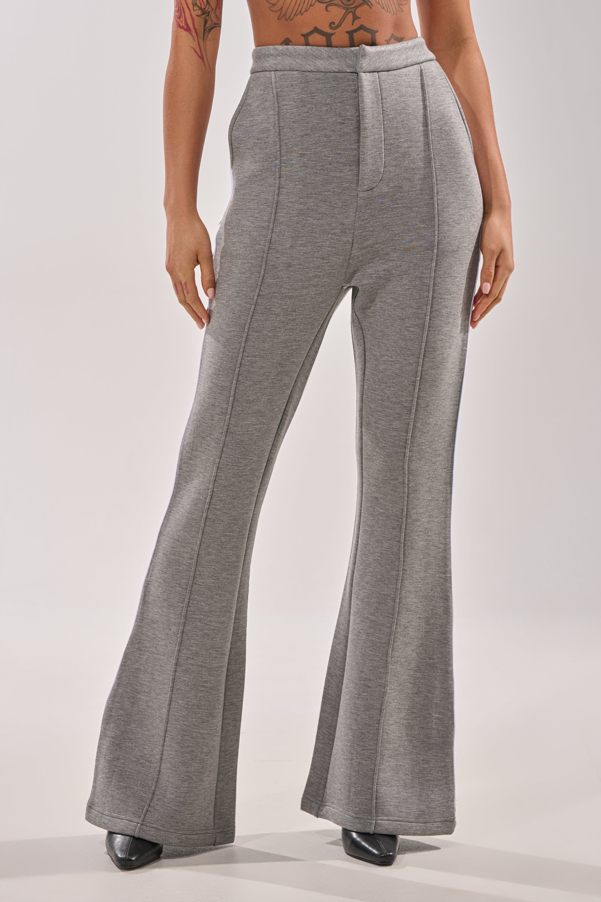 A person wears the YOU KNOW THE DRILL TROUSER—high-waisted, gray, flared-leg pants with seam details—paired with black pointed-toe shoes. The photo captures the lower torso and legs against a plain light background.
