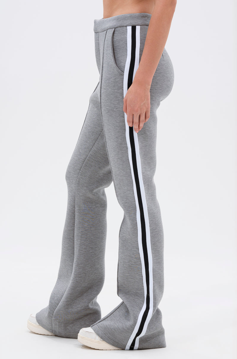 A person wears the YOU KNOW THE DRILL TROUSER—high-rise fit, grey flared athleisure pants with black and white side stripes—paired with white sneakers, set against a plain light background.