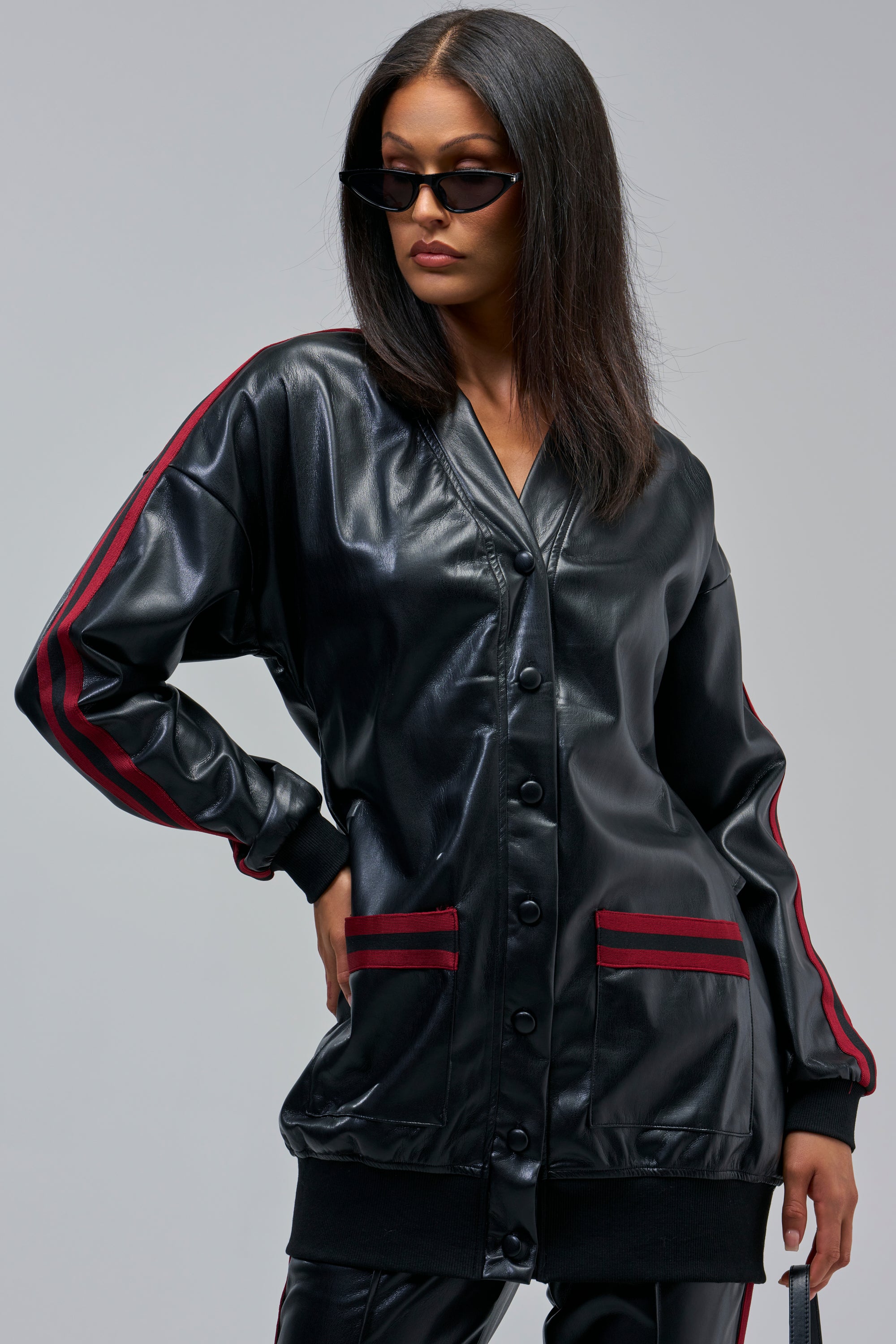 Woman wears COME AND GET IT BUTTON DOWN in black faux leather with red stripes and matching pants, oversized and longline, paired with sunglasses, posing confidently against a gray background.