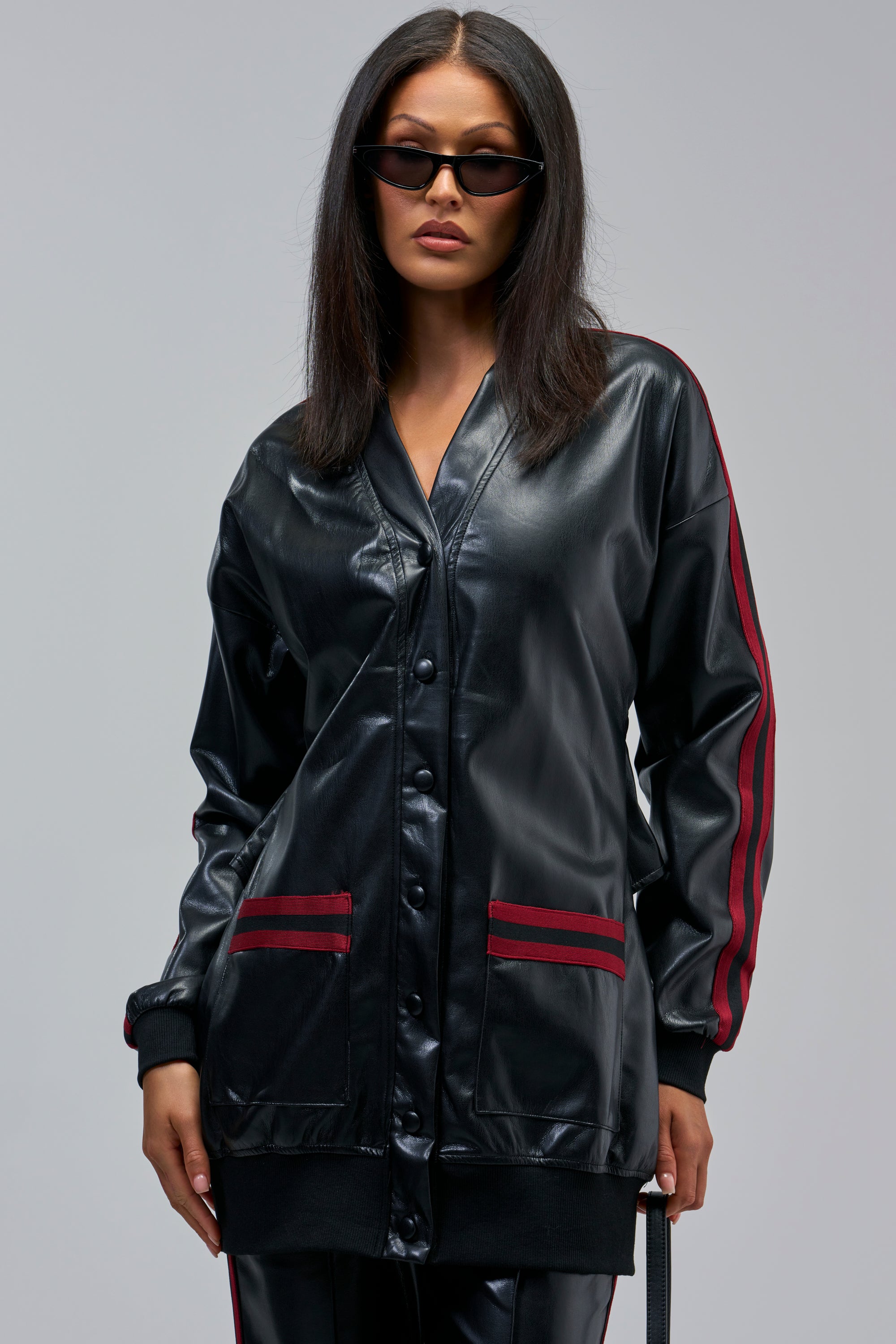 Woman wearing the COME AND GET IT BUTTON DOWN, a long black faux leather jacket with an oversized silhouette, red stripes on the sleeves, and red-trimmed pockets, styled with matching pants and sunglasses.
