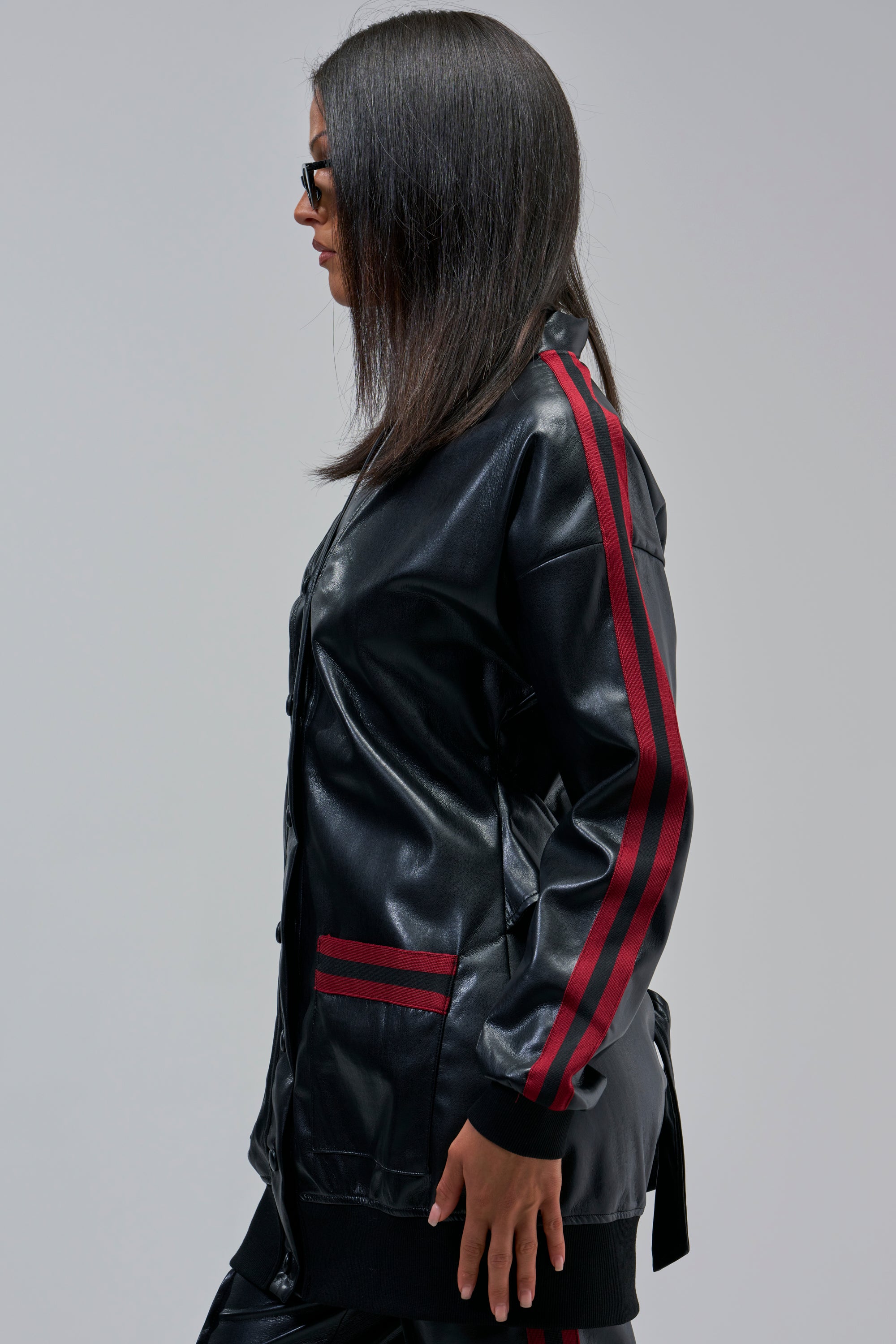 Woman with straight dark hair and sunglasses wears the COME AND GET IT BUTTON DOWN, a black faux leather jacket with red sleeve stripes and oversized longline fit, standing in profile against a light gray background.
