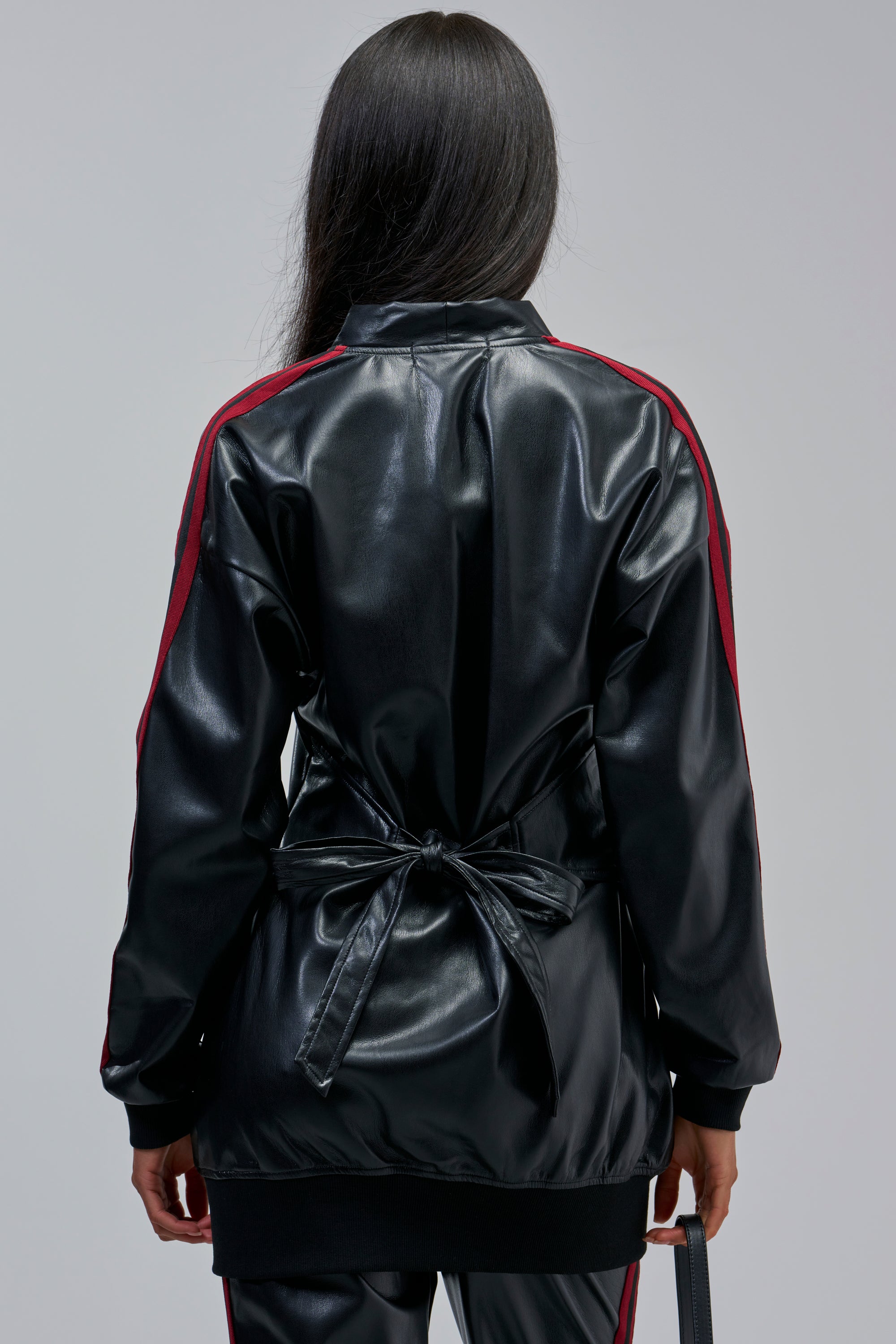 A person stands facing away, wearing the COME AND GET IT BUTTON DOWN—a shiny black faux leather top with a waist tie and red striped sleeves—paired with matching black pants.