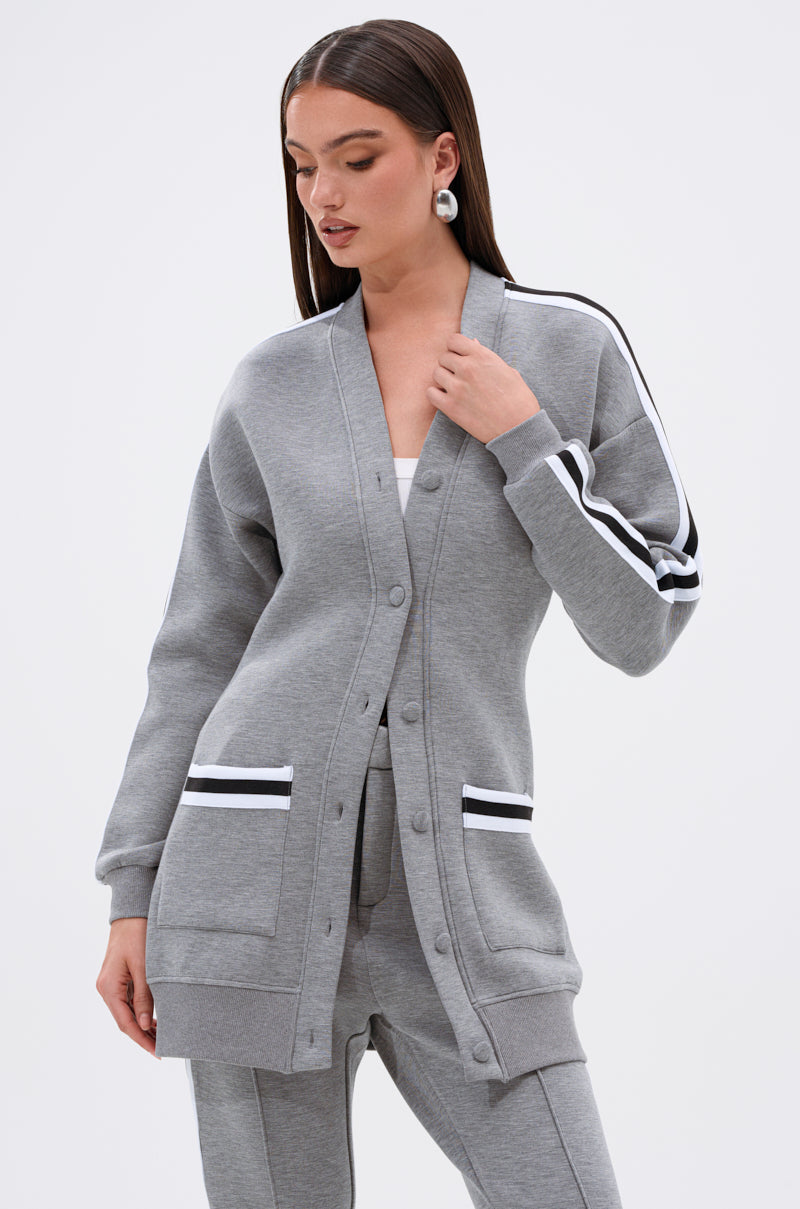 A woman stands against a plain background wearing the YOU KNOW THE DRILL CARDIGAN, a long, light gray varsity-inspired button-up with black and white striped sleeve and pocket details, layered over matching gray pants.