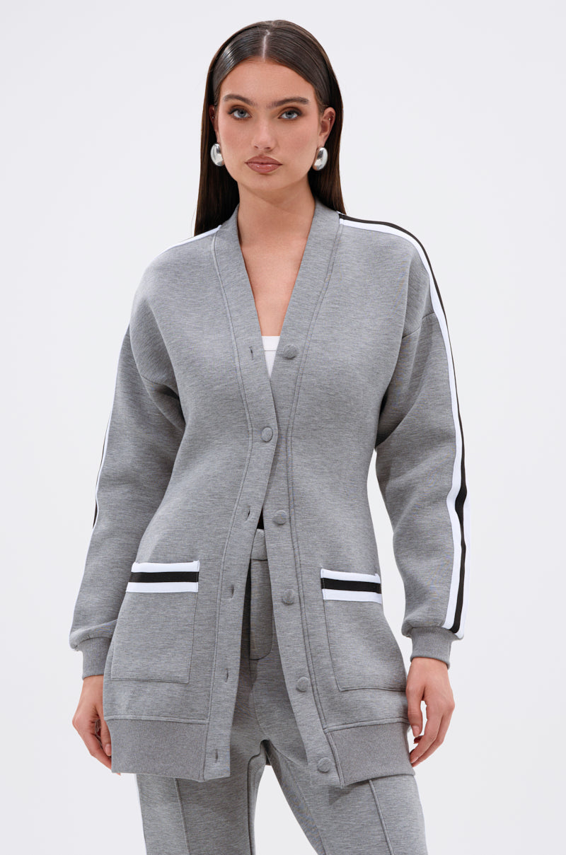 A woman with long dark hair models the YOU KNOW THE DRILL CARDIGAN, a varsity-inspired grey button-up with white and black striped sleeves, paired with matching grey pants and a white top against a light background.