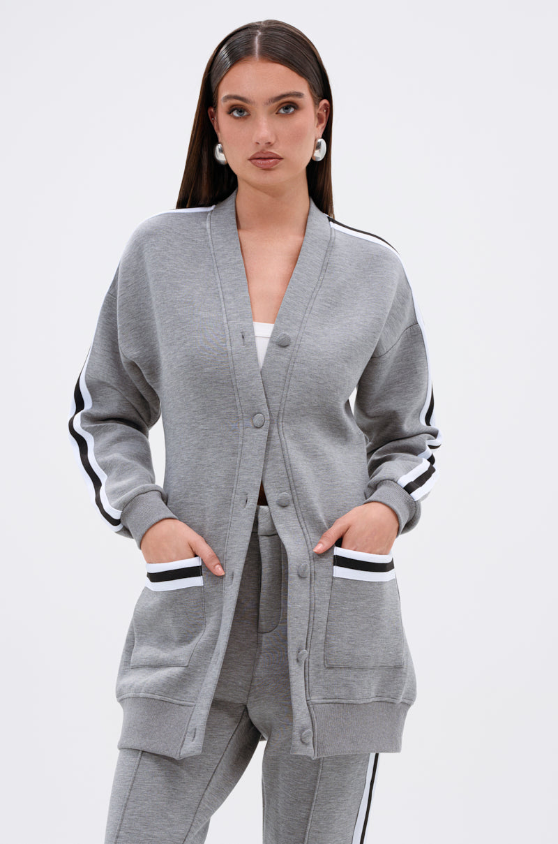 A woman with long dark hair wears the YOU KNOW THE DRILL CARDIGAN—a varsity-inspired grey button-up with black and white striped sleeves and pockets—paired with matching pants, standing against a plain white background.