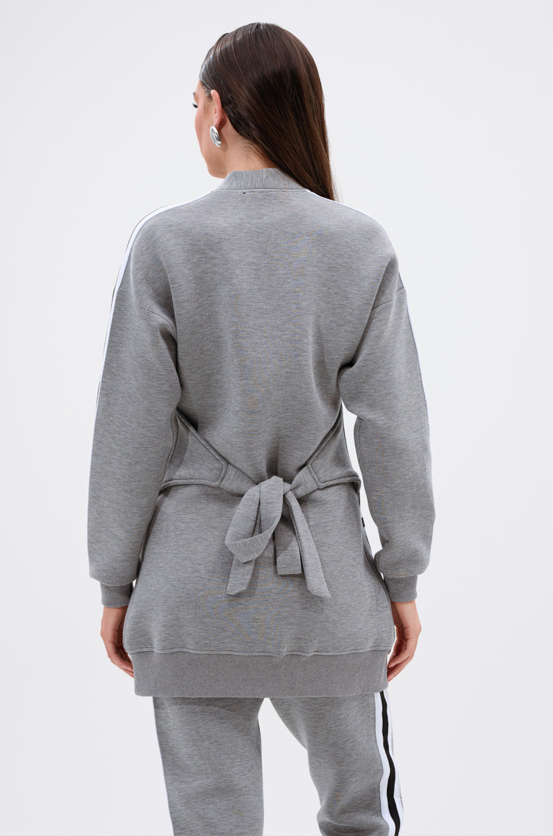 A woman with long brown hair poses from behind in the YOU KNOW THE DRILL CARDIGAN, a gray belted sweatshirt paired with matching gray pants featuring white side stripes, showcasing sporty street style against a plain light background.