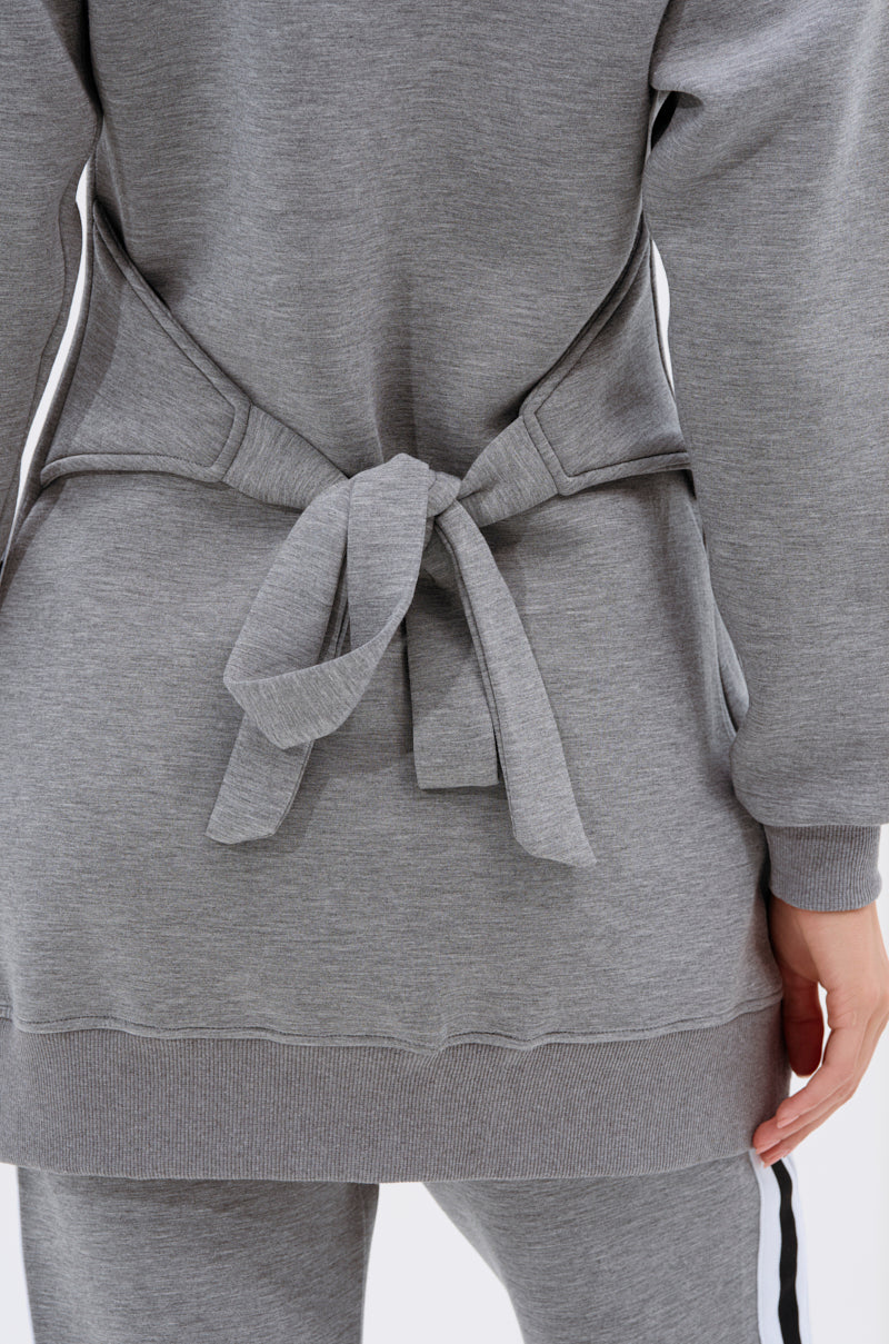 A close-up of a person wearing the YOU KNOW THE DRILL CARDIGAN in gray, varsity-inspired style with matching pants, belt tied in a bow at the waist. Only the lower torso and part of one hand are shown from the back.