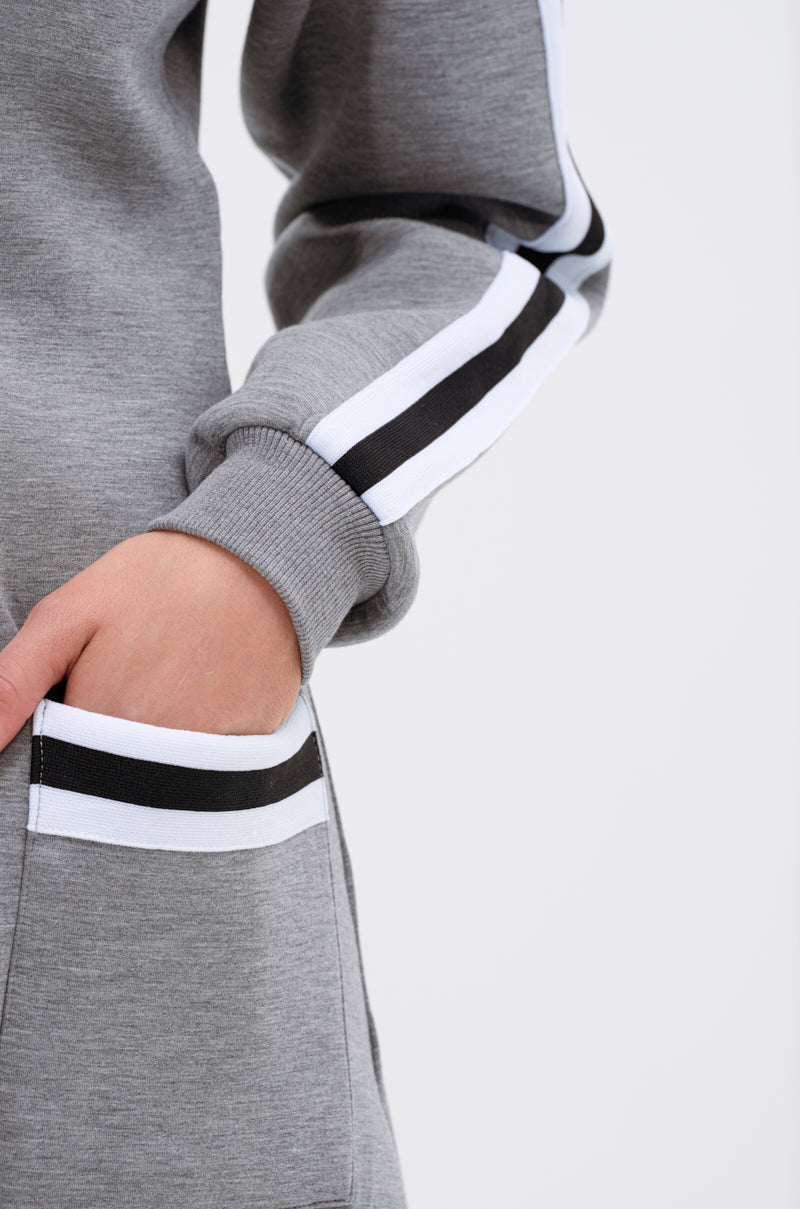 A person wearing the YOU KNOW THE DRILL CARDIGAN, a grey varsity-style piece with black and white stripes on the sleeves and pocket openings, stands with one hand partially tucked into a pocket against a plain light background.