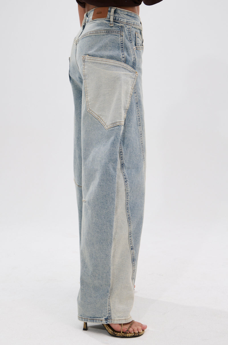 A person stands in profile on a white background, wearing the LOTS TO GIVE BARREL JEANS with oversized patch pockets and light wash, paired with open-toe high-heeled sandals.