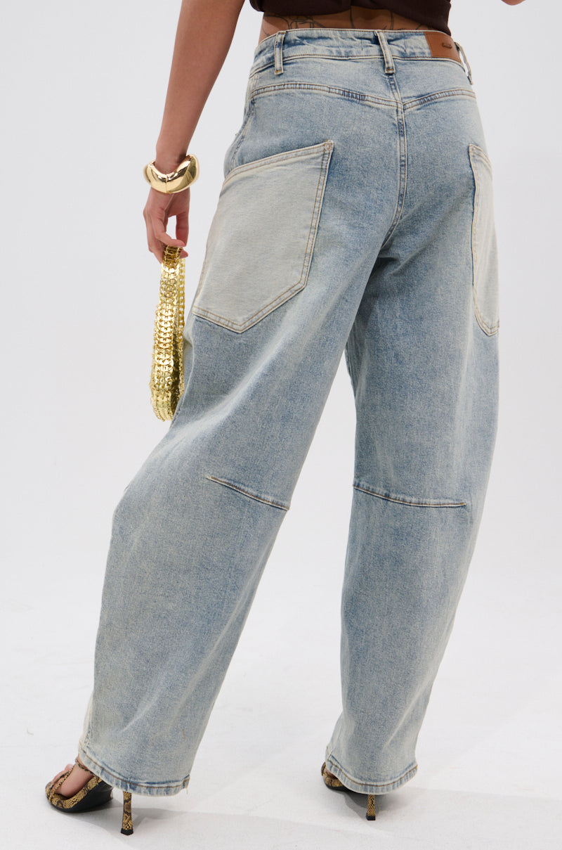 Wearing LOTS TO GIVE BARREL JEANS in a light wash with large back pockets, a person pairs them with a brown top, black open-toe heels, gold accessories, and a shiny gold textured clutch. The photo is taken from behind.