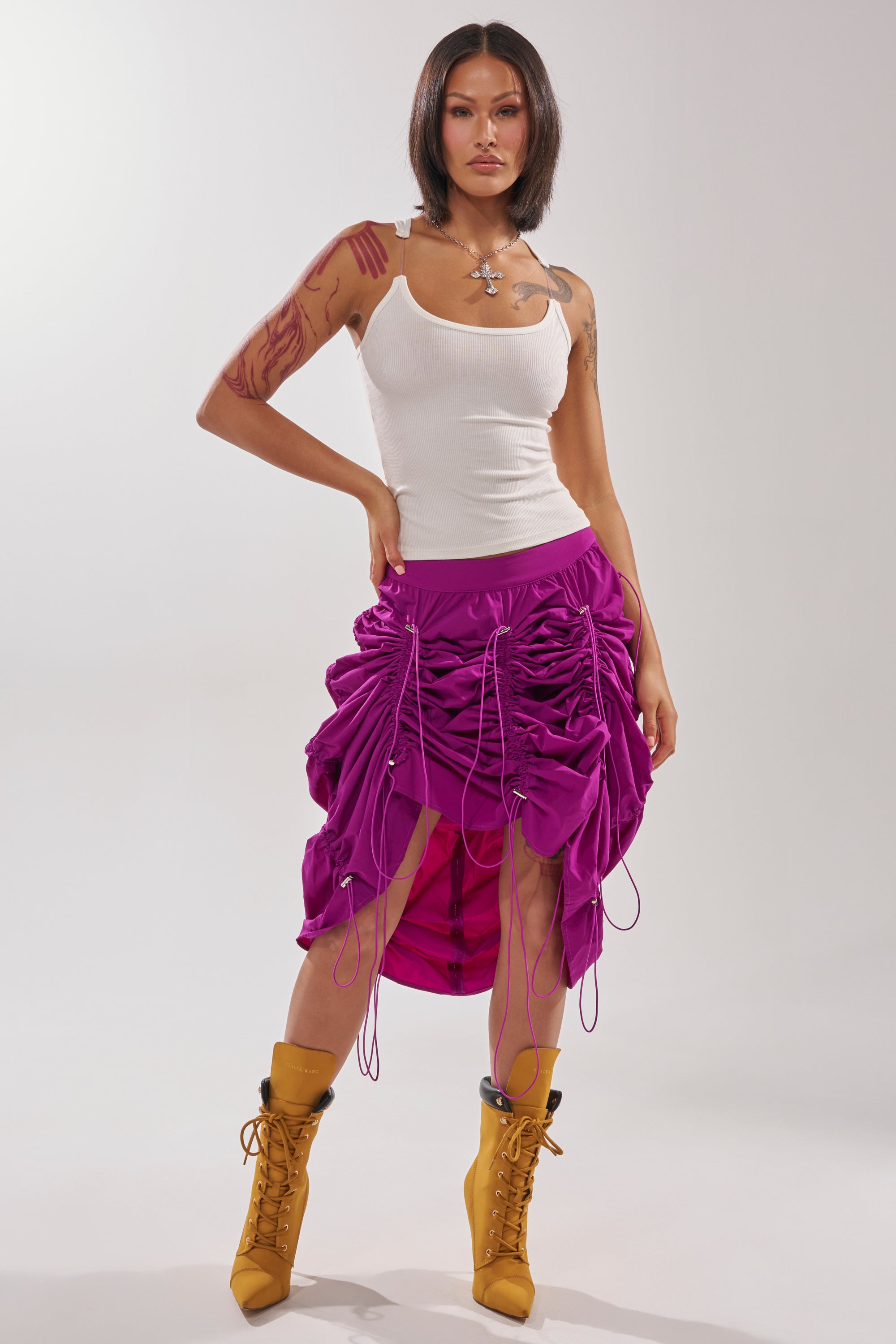 A woman with tattooed arms wears a white tank top, the BELLA PARACHUTE MAXI SKIRT IN FUCHSIA, and yellow lace-up boots as she stands confidently against a plain light background. 9