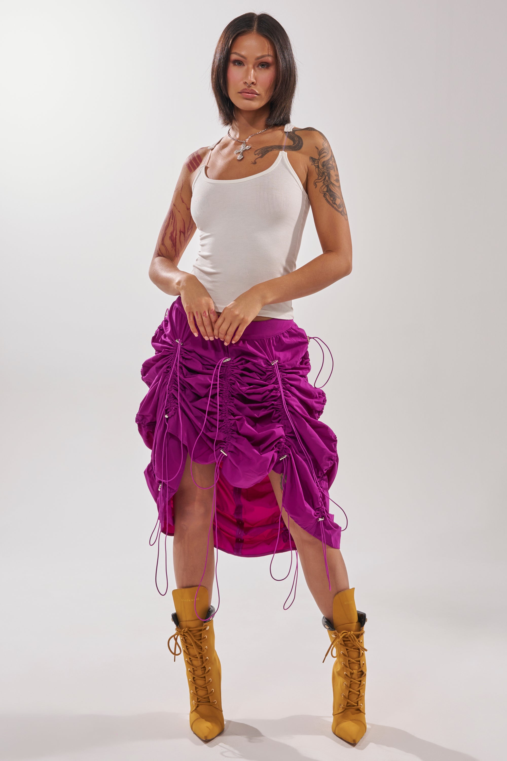 A woman with tattoos poses in a white tank top, mustard yellow lace-up boots, and the BELLA PARACHUTE MAXI SKIRT IN FUCHSIA with adjustable drawstrings, standing against a plain light background. 10