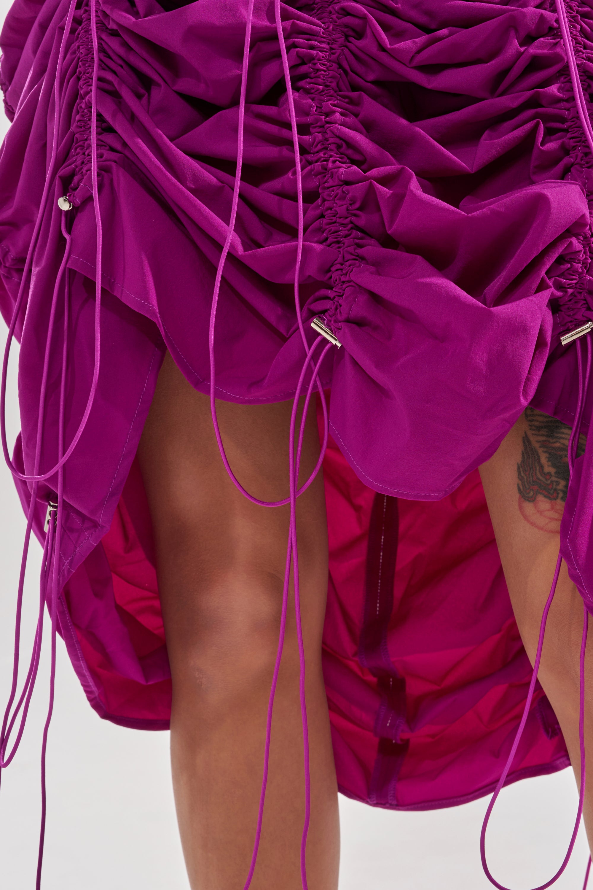 A close-up of a person wearing the BELLA PARACHUTE MAXI SKIRT IN FUCHSIA, featuring ruched fabric, drawstrings, and a high-low hem that reveals their legs and part of a thigh tattoo. 12
