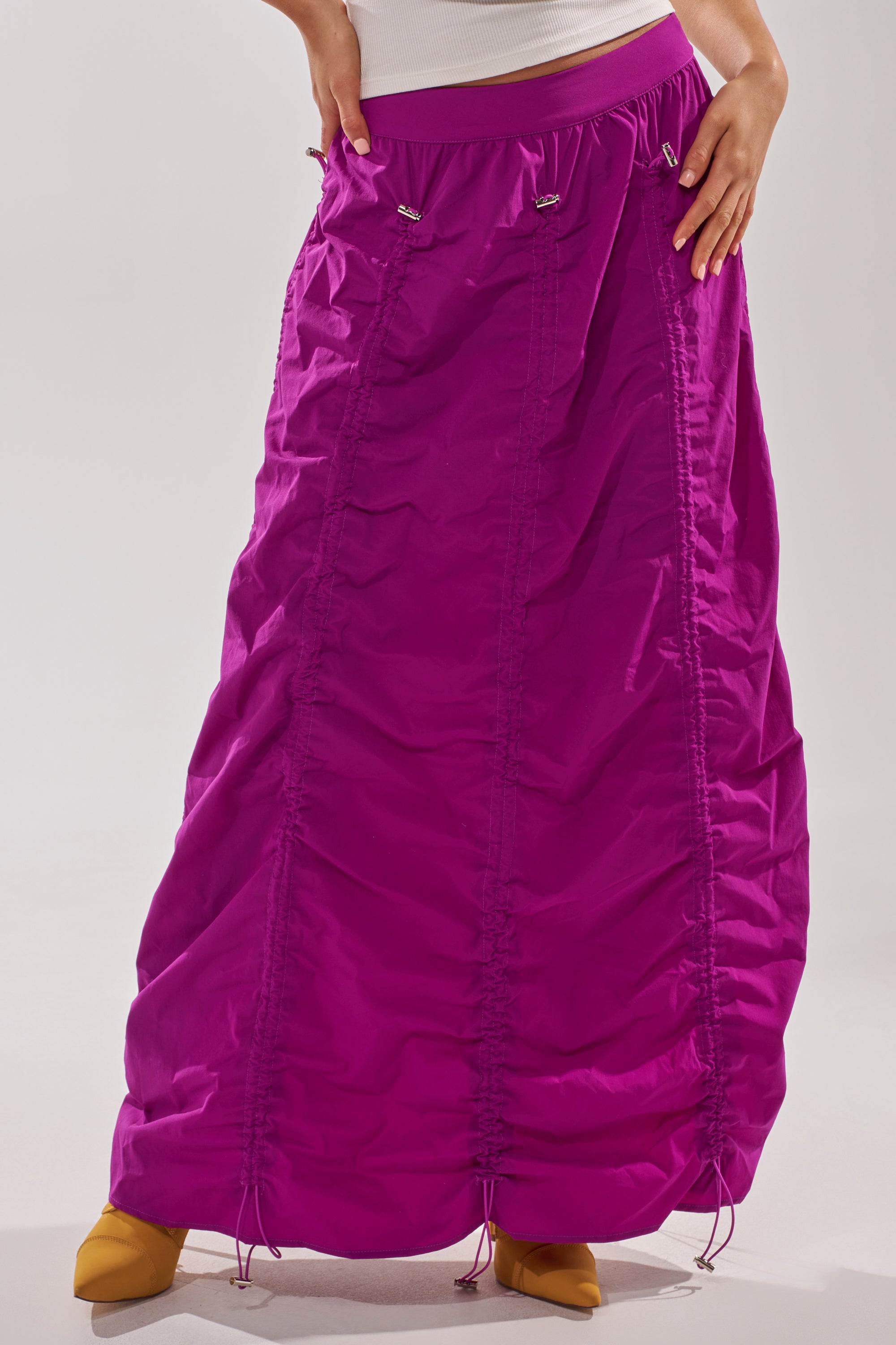 A person wears the BELLA PARACHUTE MAXI SKIRT IN FUCHSIA, featuring adjustable drawstrings, paired with yellow pointed-toe shoes. Only the lower half of their body is visible. 3