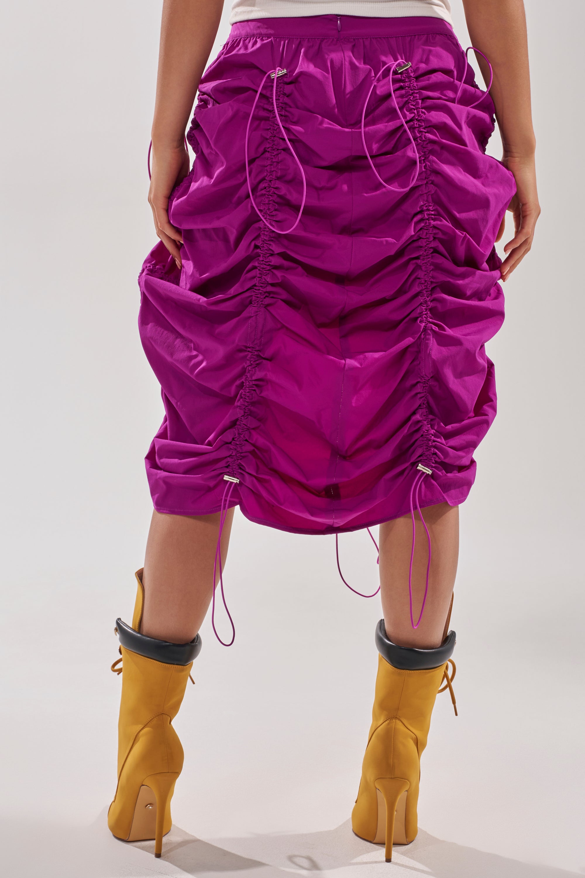Model wears the BELLA PARACHUTE MAXI SKIRT IN FUCHSIA with adjustable drawstrings and yellow high-heeled boots, shown from the back against a neutral backdrop. 6