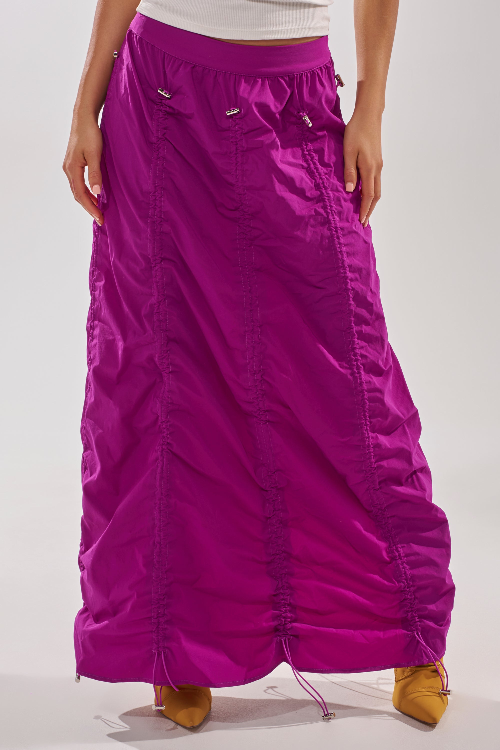 Only the lower body is visible against a plain background, featuring the BELLA PARACHUTE MAXI SKIRT IN FUCHSIA with ruched drawstring details, styled with yellow pointed-toe shoes. 7